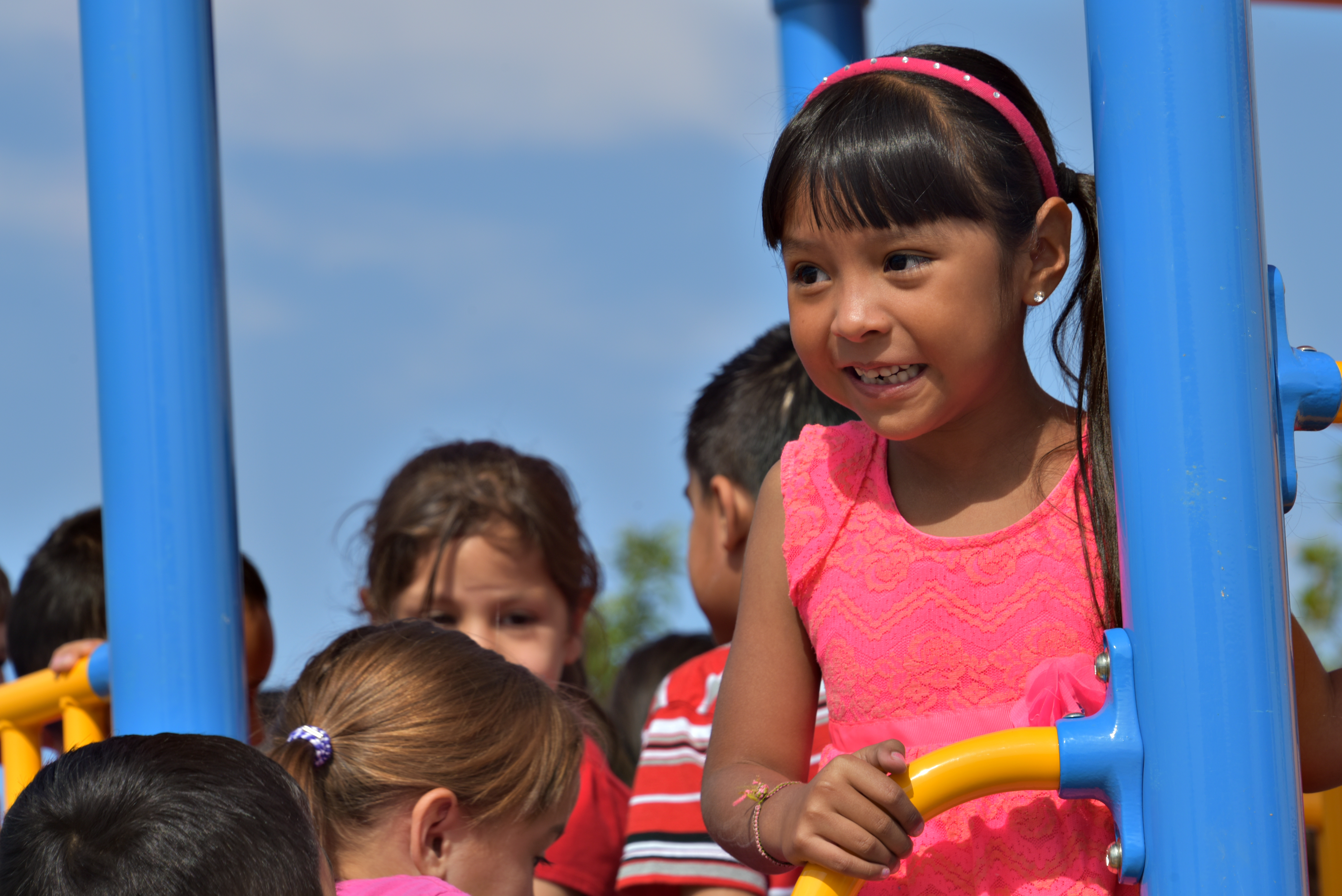 A little girl in a pink dress smiles on the playground surrounded by other kids